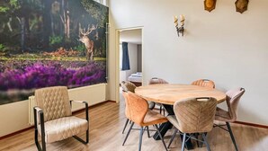 A dining area with a round wooden table and cushioned chairs, a large forest-themed mural on the wall, and two mounted deer heads. - Cosy forest bungalow with sauna (Kootwijk)