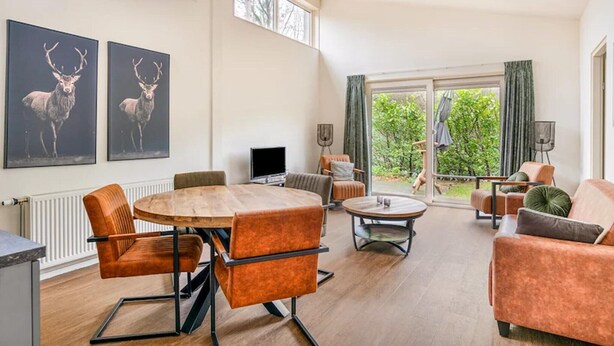 A spacious living area with a round wooden table, orange upholstered chairs, a sofa, and a television. There are two large windows and a view of greenery outside. - Cosy forest bungalow with sauna (Kootwijk)
