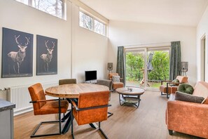 Un salón amplio con una mesa redonda de madera, sillas tapizadas de color naranja, un sofá y un televisor. Hay dos ventanas grandes y vistas al verdor exterior.