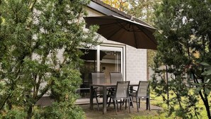 An outdoor patio with a table and chairs, a large umbrella, and a building with a glass door. - Cosy forest bungalow with sauna (Kootwijk)