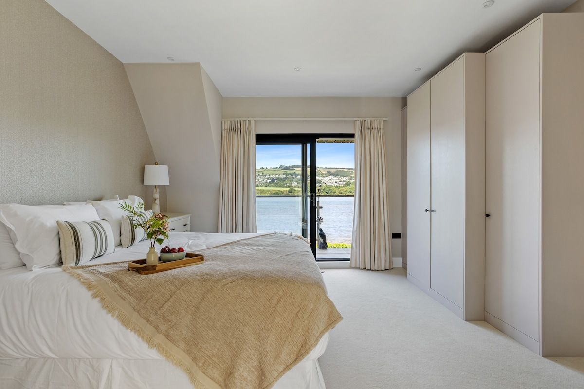 A bedroom with a large bed, a bedside table with a lamp, a view of the outdoors through a sliding door, and a wardrobe.