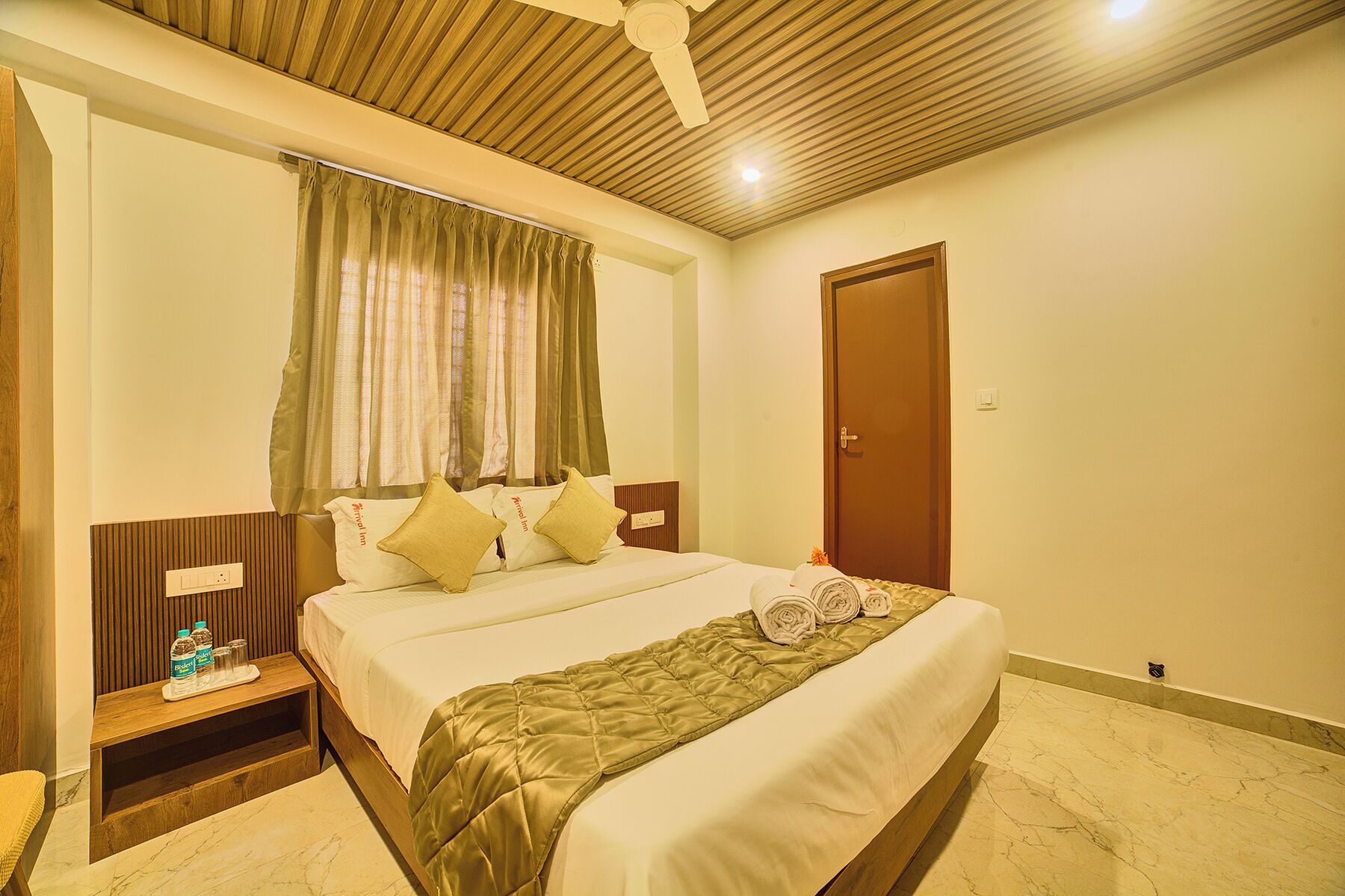 A hotel room with a bed, a bedside table with bottles, a wooden headboard, a ceiling fan, and a door.