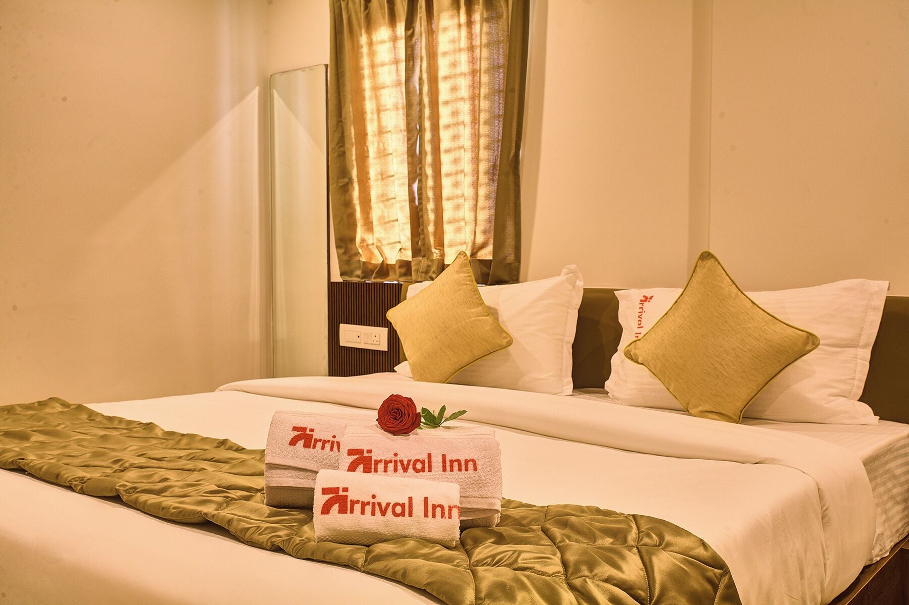 A neatly made bed with a green bedspread and pillows, a bedside table with a lamp, and a window with curtains.