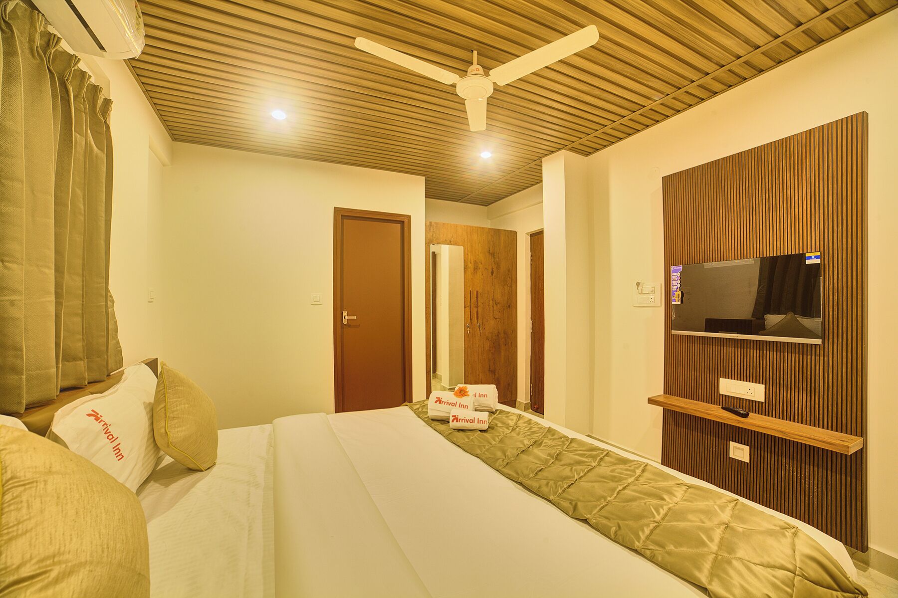 A hotel room with a bed, a ceiling fan, a TV mounted on a wooden panel wall, and a door leading to another room.