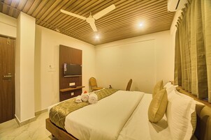 Habitación de hotel con una cama grande, un televisor de pantalla plana montado en un panel de madera, un ventilador de techo y una ventana con cortinas.