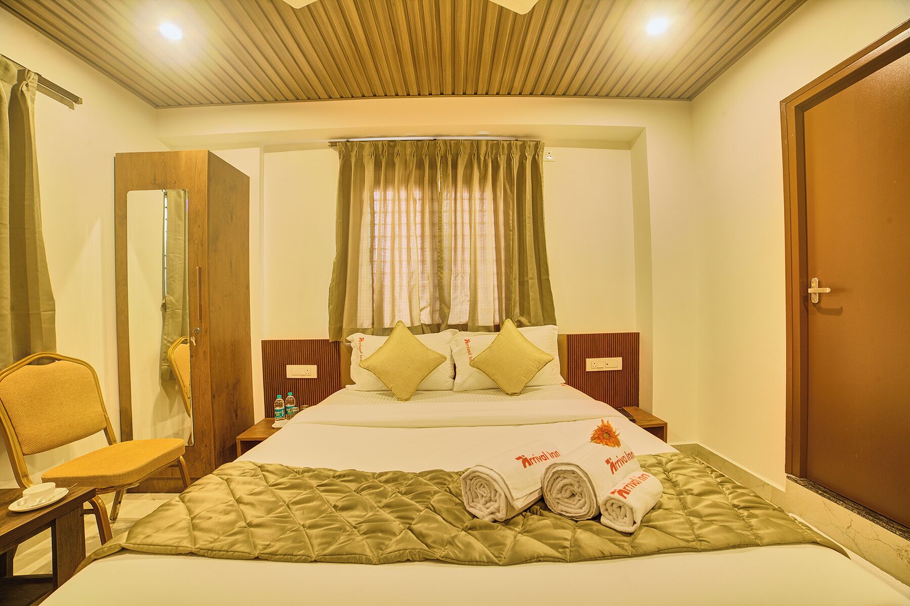 A hotel room with a bed, a chair, a wooden headboard, a window with curtains, and a door.