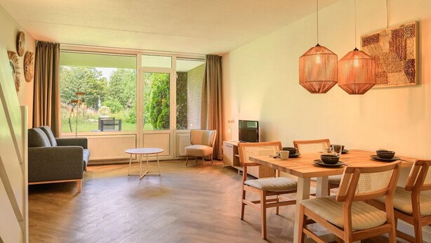 A cozy living area with a sofa, a small round table, and a dining table with chairs. There is a large window providing a view of the outdoors. - Jungle-themed family escape (Bruinisse)