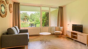A modern living room with a sofa, a small round table, a television on a wooden cabinet, and large windows offering a view of greenery outside. - Jungle-themed family escape (Bruinisse)