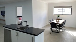 Una cocina moderna con encimera negra y electrodomésticos de acero inoxidable, un comedor con mesa y sillas, y un amplio ventanal que aporta luz natural.