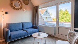 A modern living room with a blue sofa, a round wooden coffee table, and a view of a house through large sliding glass doors. - Modern apartment near centre facilities (Bruinisse)