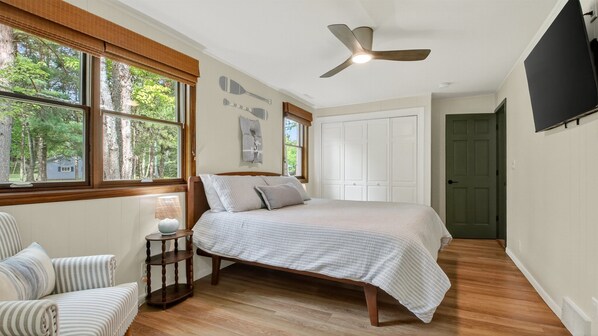 A bedroom with a bed, a nightstand, a chair, a ceiling fan, and a window with a view of trees.