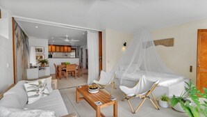 A modern living room with a sofa, a wooden coffee table, and a canopy bed. There is a dining area with a table and chairs, and a kitchen in the background.