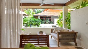 A room with a wooden bench, a sofa, and a table with a plant. There is a window with a view of greenery and a building with a tiled roof.