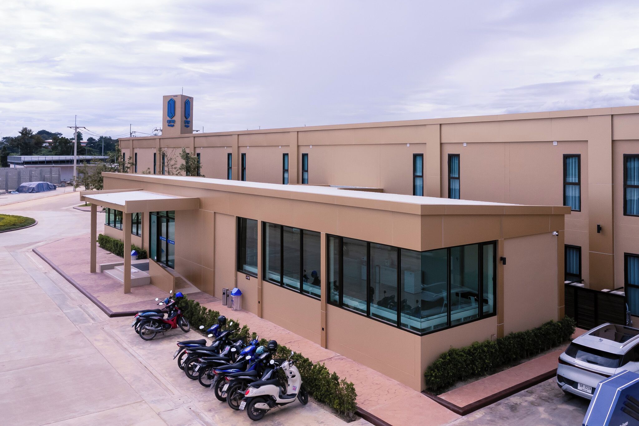 A modern building with a flat roof, large windows, and a covered entrance area. There are several parked scooters and a car near the entrance.