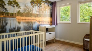 Une pièce décorée d’une fresque représentant un lac et des arbres, avec un berceau, une commode blanche et un fauteuil en osier.