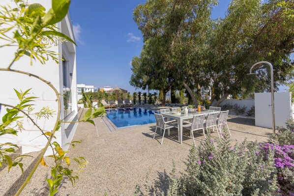 A swimming pool with lounge chairs and a table, surrounded by greenery and a white building.