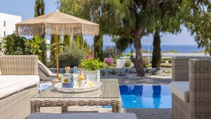 A poolside setting with wicker furniture, a table with a tray of drinks and a plant, and a thatched umbrella. - Protaras Leomi Seaview Villa (Paralimni)