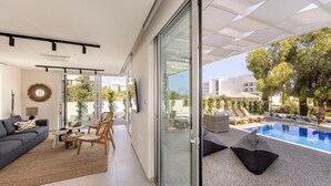A modern living room with a sofa, chairs, and a coffee table. There is a sliding glass door leading to a pool area with lounge chairs and a pergola. - Protaras Leomi Seaview Villa (Paralimni)