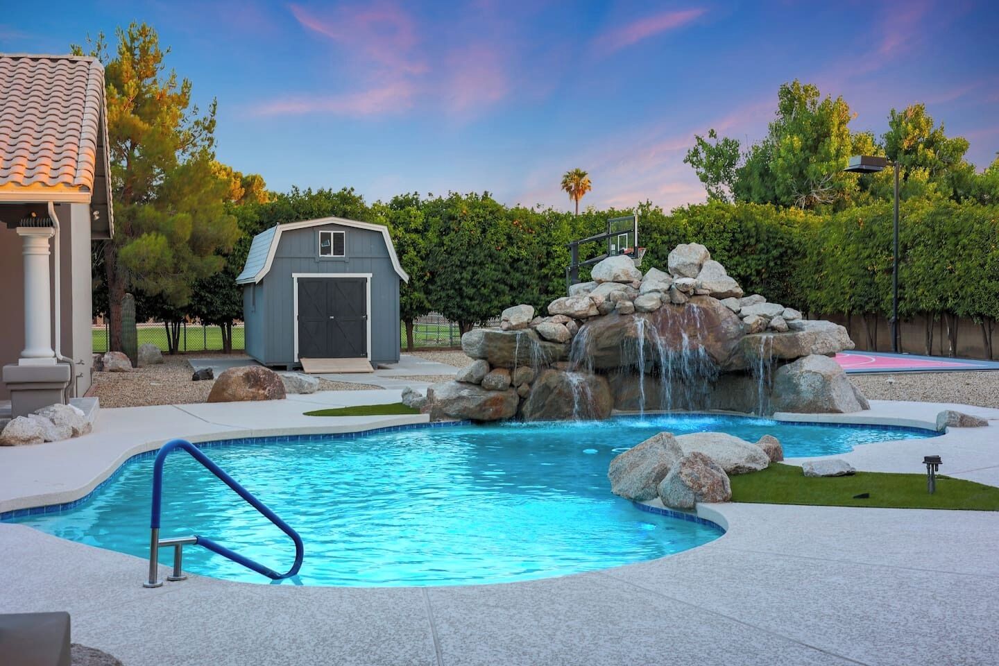 A swimming pool with a waterfall feature, a covered storage shed, and a basketball court in the background.