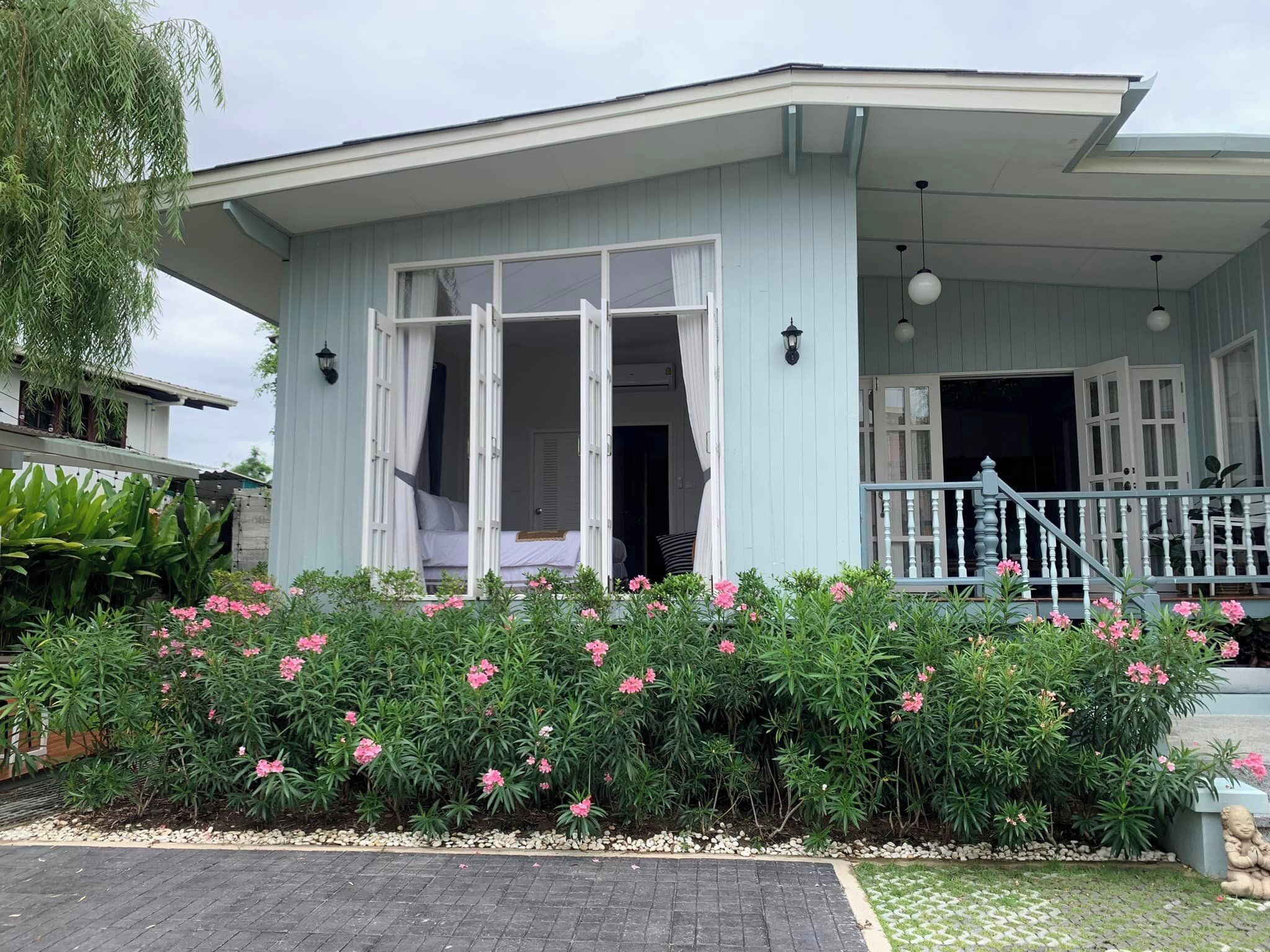 A light blue building with a covered porch, a bed visible inside, and a garden with pink flowers in front.