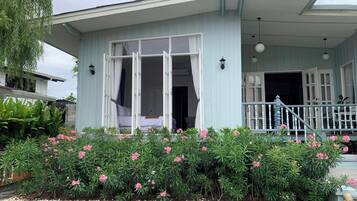 A light blue building with a covered porch, a bed visible inside, and a garden with pink flowers in front.