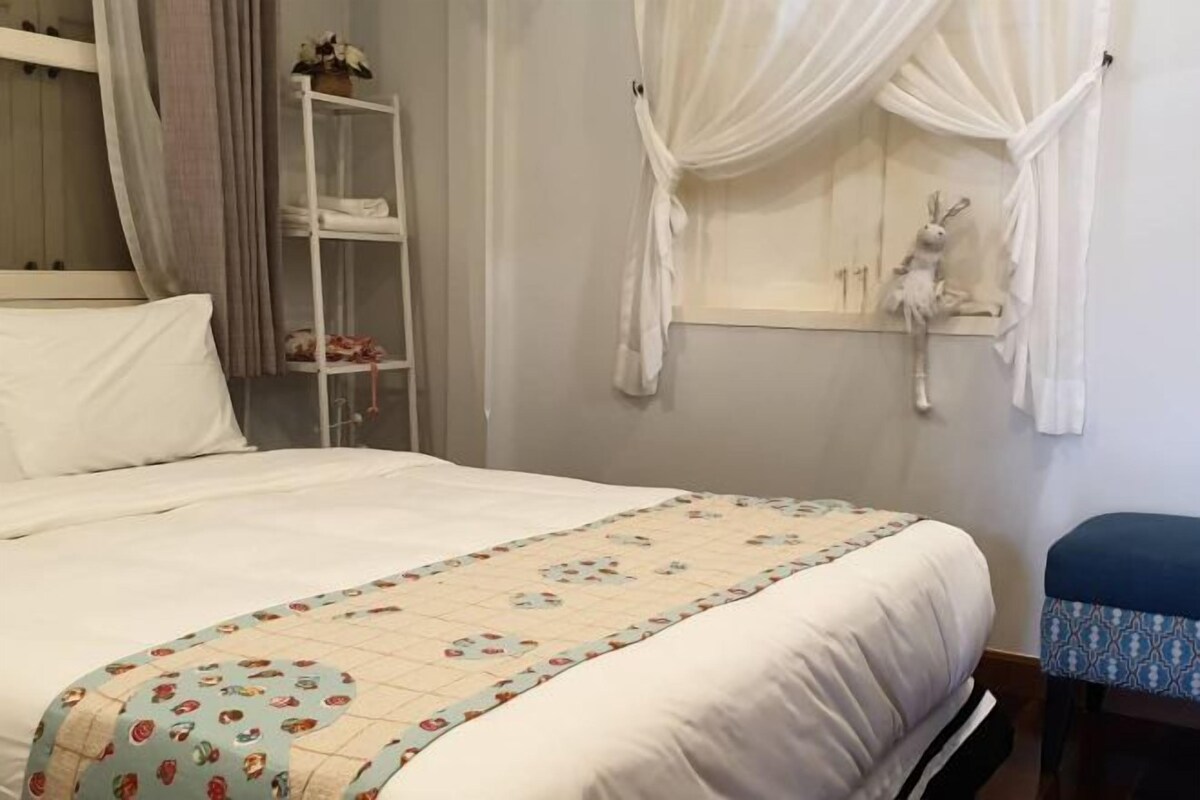 Una cama bien hecha con una colcha de motivos florales, una pared blanca con un ventanal y un estante blanco con toallas dobladas.