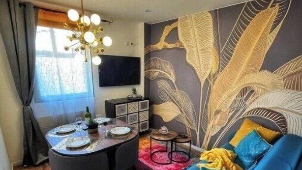 Un salon moderne avec un canapé bleu, une table à manger dressée pour quatre personnes et un mur décoratif orné de motifs de feuilles.
