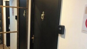 Une porte en bois sombre, dotée d’une poignée en laiton et d’une petite plaque en laiton, un grand miroir avec un cadre en bois, et un interrupteur sur le mur.