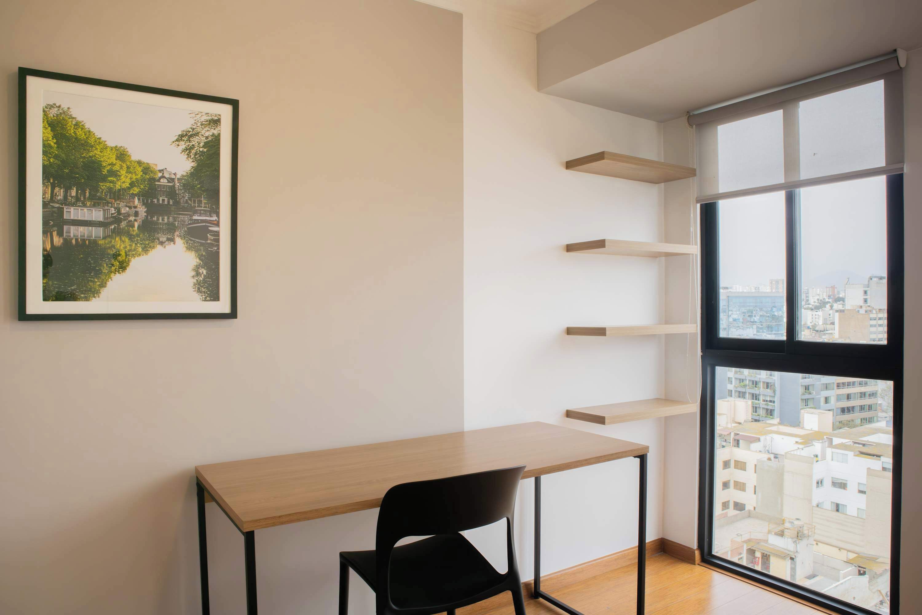 A minimalist room with a wooden desk, black chair, and a cityscape view through a large window. Shelves are mounted on the wall.