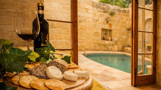 Ein Holzboden mit Käse, Crackern und einer Weinflasche, vor einer Steinsäule und mit Blick auf einen Pool.