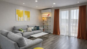 A modern living room with grey sofas, a yellow stool, and a bookshelf with a sign reading "AIR MALTA".