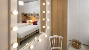 A bedroom with a large mirror surrounded by lights, two beds, and a chair.