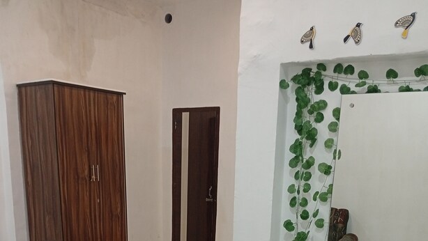 A room with a wooden wardrobe, a dining table with chairs, a mirror with a green plant border, and a door. - BLOOM STAY  (Amritsar)