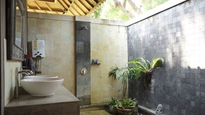 Une salle de bain avec un grand lavabo blanc, une douche et un mur en pierre. On y trouve des plantes et une petite statue accrochée au mur.