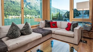 Uma sala de estar moderna com um amplo sofá de canto, uma mesa de centro de vidro e vista para um lago e montanhas através de grandes janelas.