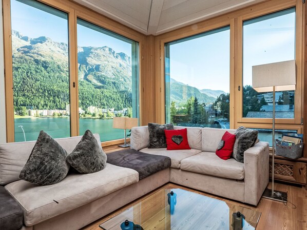A modern living room with a large sectional sofa, a glass coffee table, and a view of a lake and mountains through large windows. - Chesa Spuonda Verde 2.5 by Interhome (St. Moritz)