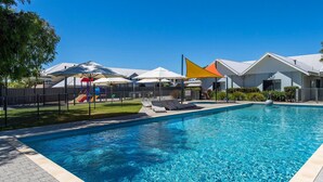 Une piscine avec des chaises longues et des parasols, une aire de jeux clôturée et un bâtiment doté d’un toit blanc.