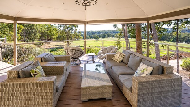 A covered outdoor seating area with wicker furniture, a glass coffee table, and a view of a green landscape. - Valley View – Ferguson Valley (Ferguson)