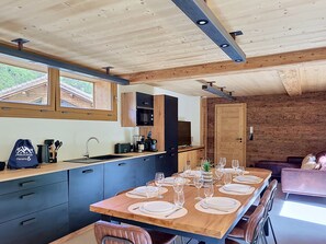 A modern kitchen with a wooden table set for dining, a built-in oven, and a view of the outdoors through large windows.