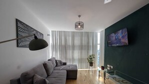Una sala de estar moderna con un sofá modular, una lámpara colgante, un televisor montado en la pared y un ventanal con persianas verticales.