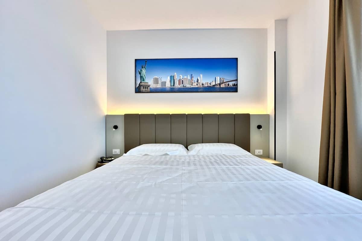 A hotel room with a large bed, a cityscape painting above it, and a nightstand with a phone.