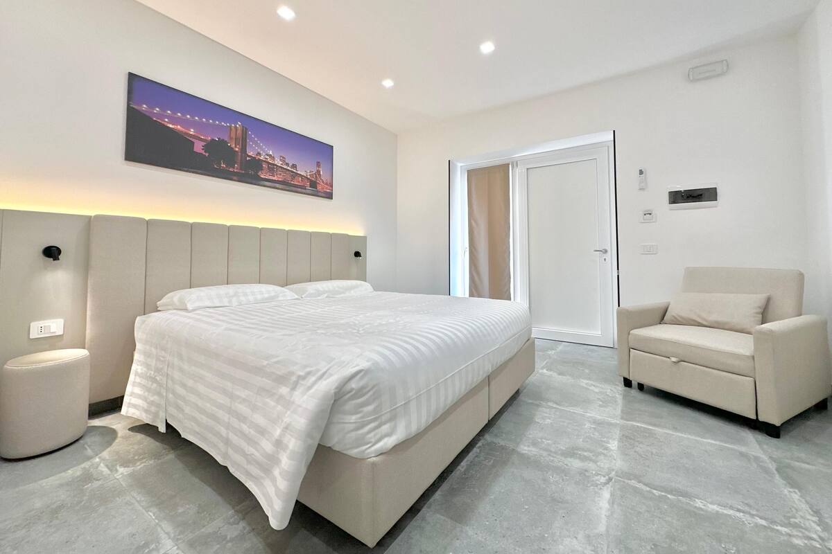 A modern hotel room with a large bed, a chair, a nightstand, and a cityscape painting on the wall.