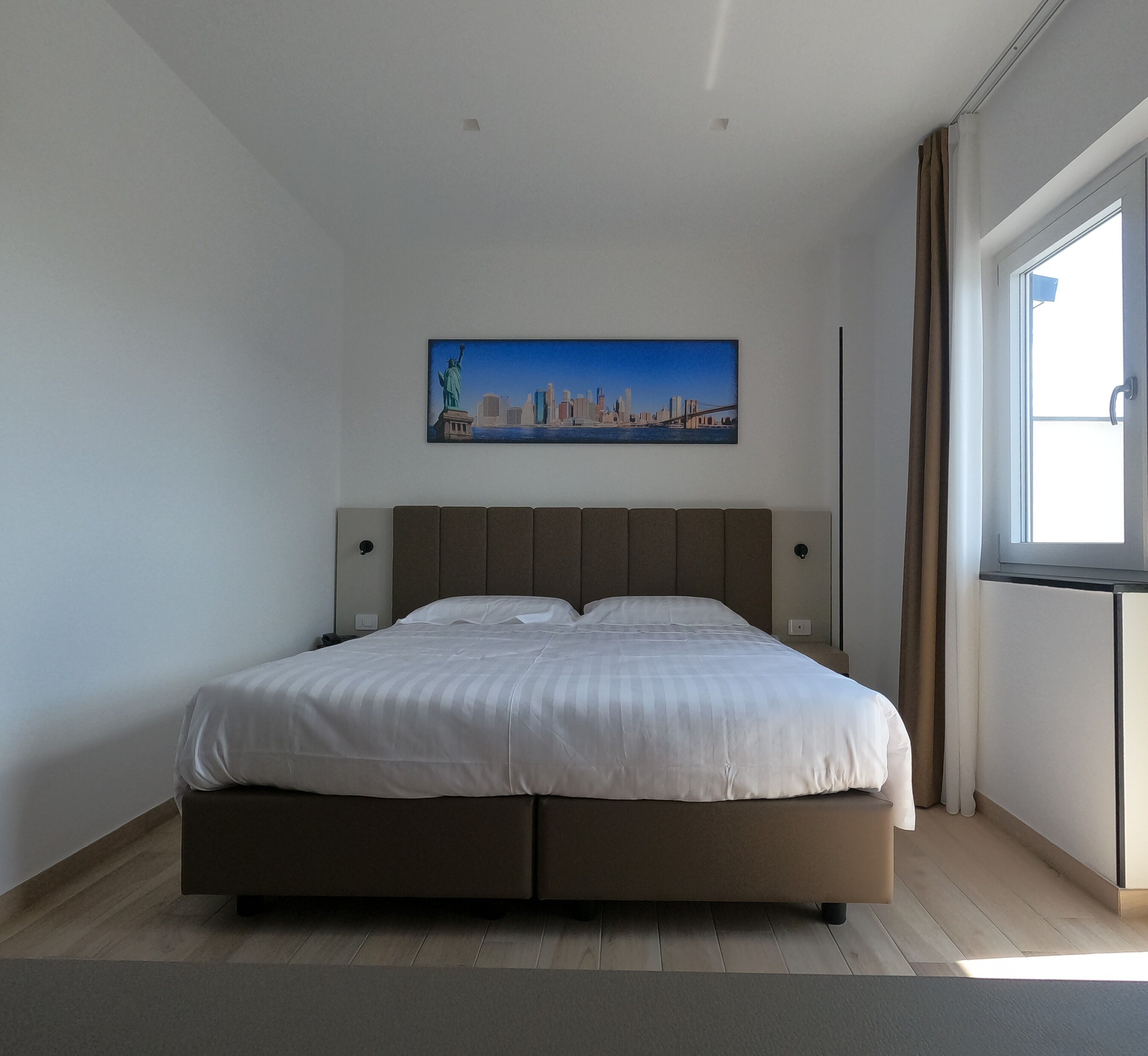 A modern bedroom with a large bed, a cityscape painting on the wall, and a window with curtains.