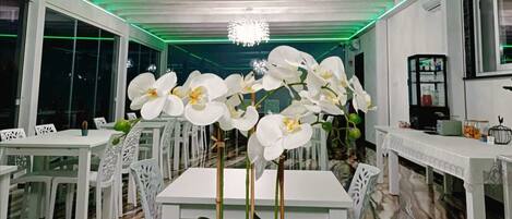 A modern dining area with white tables and chairs, a central orchid plant, and a chandelier.
