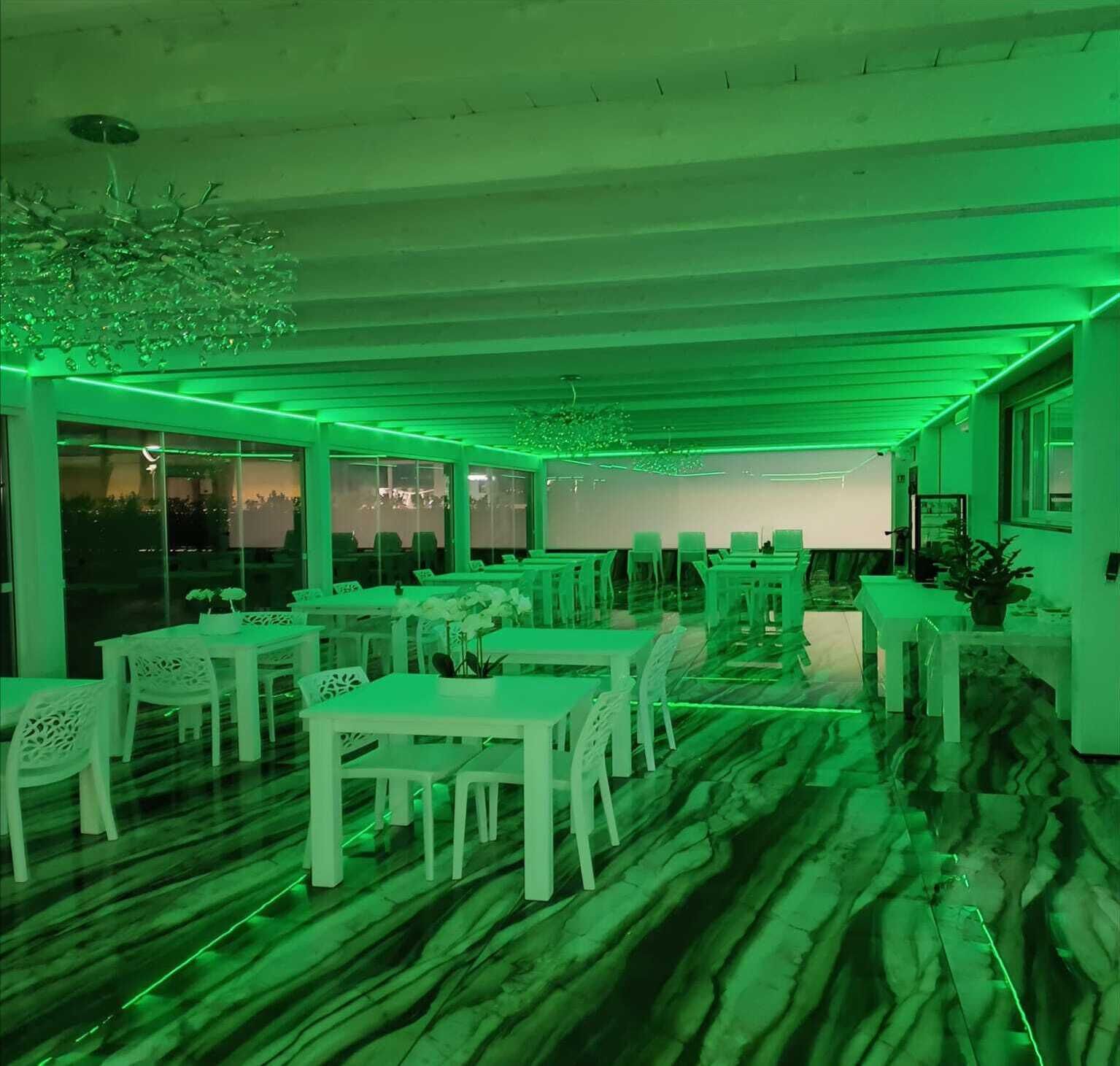 A restaurant with white tables and chairs, green ambient lighting, and a view of the outdoors through large windows.