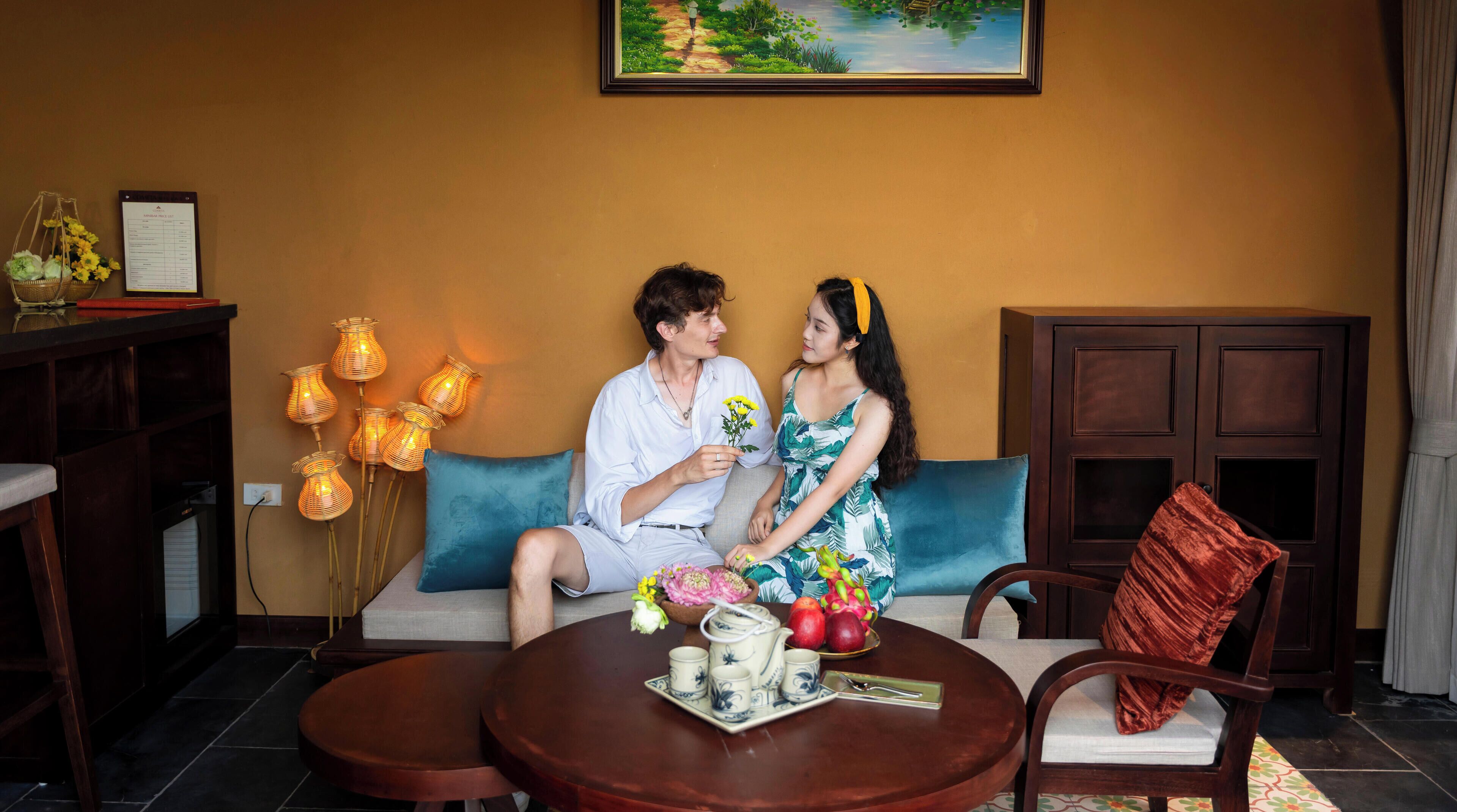 Un couple est assis sur un canapé, devant lequel se trouve une table. On aperçoit également un meuble et un tableau accroché au mur.