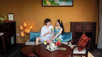 Un couple est assis sur un canapé, devant lequel se trouve une table. On aperçoit également un meuble et un tableau accroché au mur.