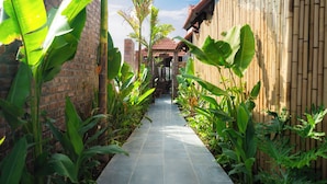 A narrow paved pathway flanked by lush green plants and a bamboo fence, leading to a brick building with a tiled roof. - Cometa Villas Ninh Bình (Gia Phong)