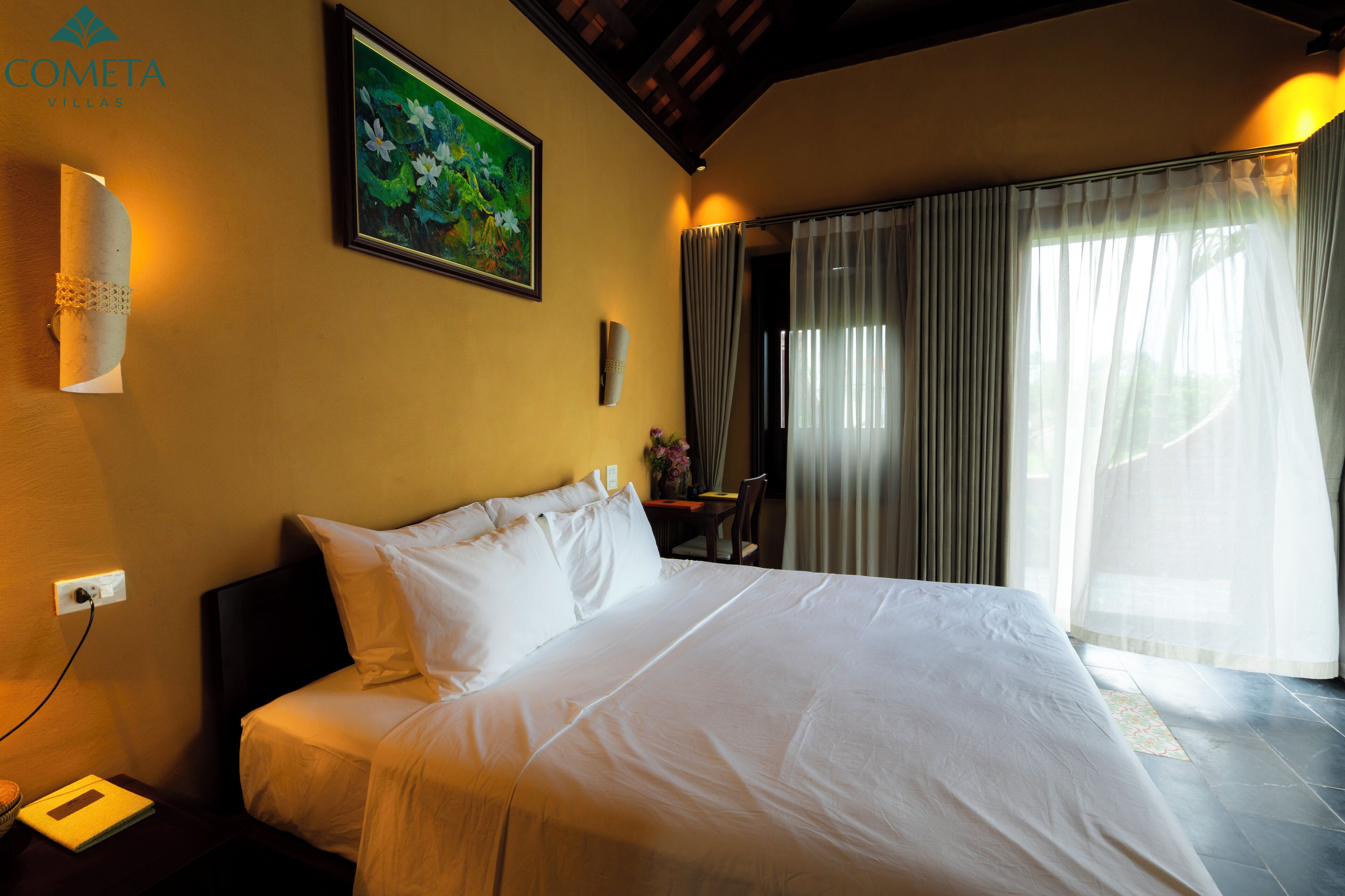 A hotel room with a large bed, a painting on the wall, a desk with a chair, and a window with curtains.