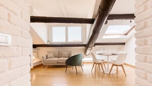 Un loft moderno con vigas de madera a la vista, una cama en el entrepiso, una zona de comedor con sillas blancas y un amplio ventanal que proporciona luz natural.